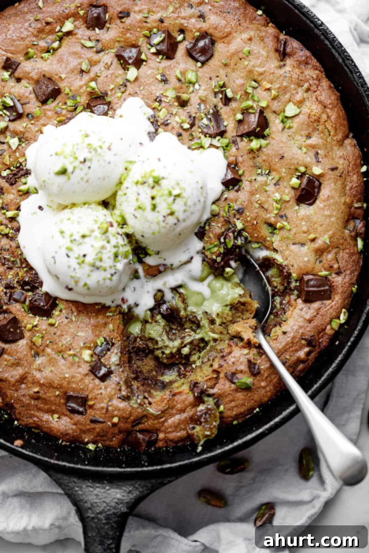 Pistachio Chocolate Chip Skillet Cookie 2 Close-up image of a spoon lifting a portion of warm pistachio cookie from a cast-iron skillet, generously served with melting vanilla ice cream and a delicate chocolate drizzle.