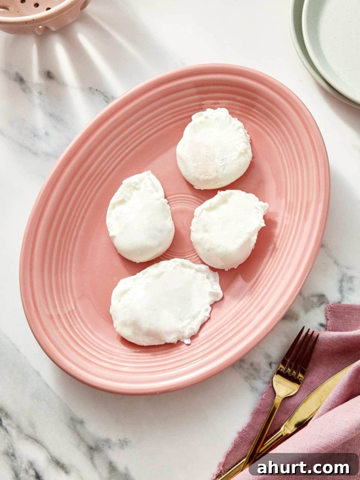 Mastering Perfect Poached Eggs 6 Four perfectly poached eggs resting on a blush-pink plate over a marble background.