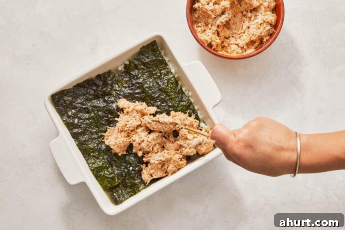 Easy At-Home Sushi Bake 8 Spoon over a dish of food, preparing to add nori and a creamy seafood topping to the rice.