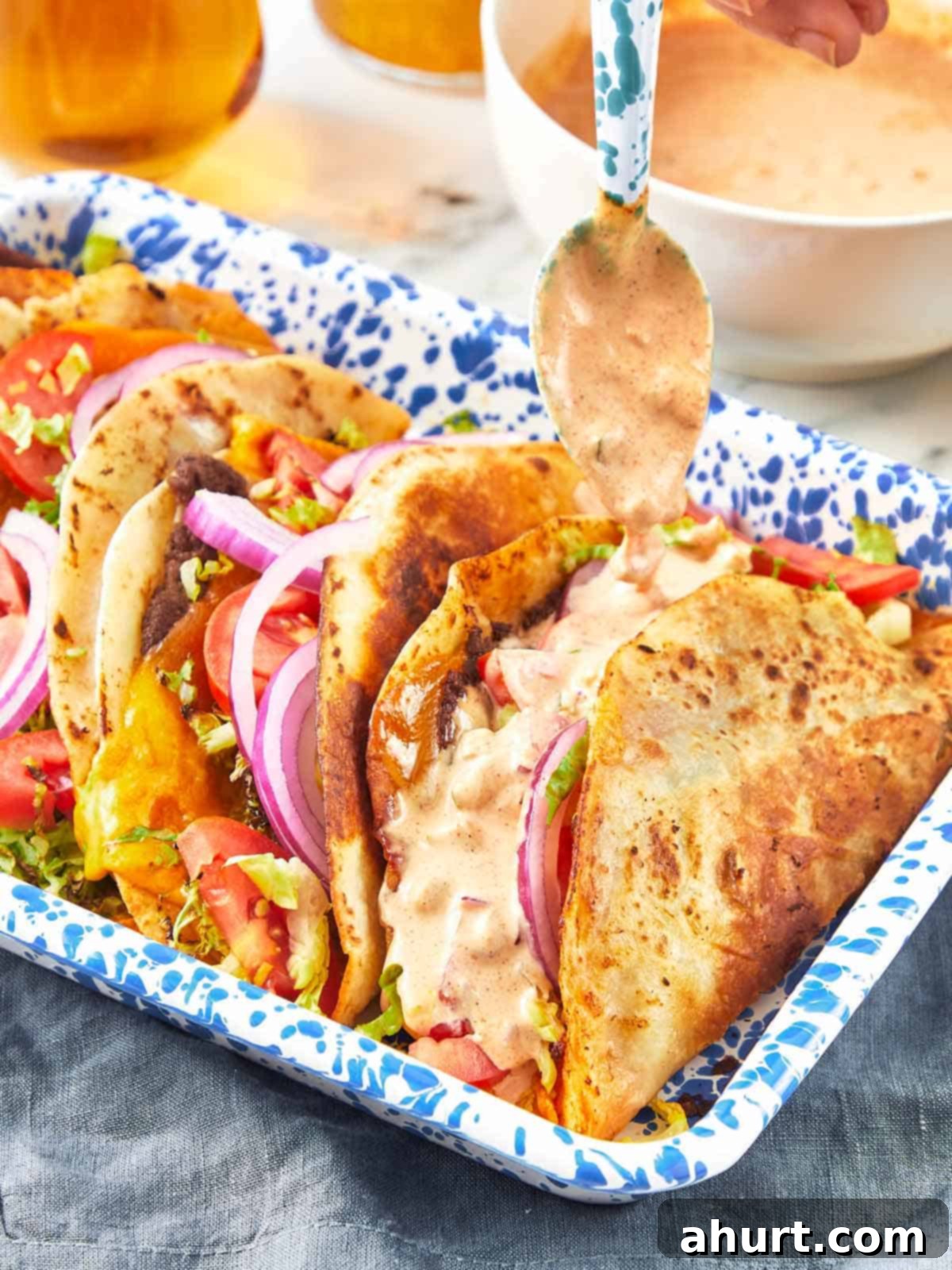 Smash Burger Taco Mashup 10 Close-up of smash burger tacos in a blue speckled tray, topped with lettuce, tomato, red onion, melted cheese, and drizzled with creamy sauce, highlighting the crispy edges.