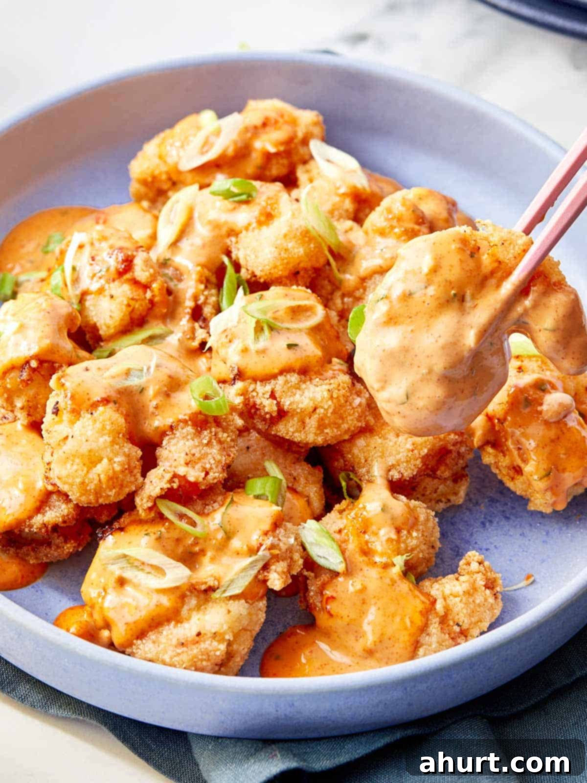 Dynamite Shrimp 10 Delicious Bang Bang Shrimp coated in creamy, spicy sauce, garnished with vibrant sliced green onions, served in an elegant blue bowl with chopsticks lifting a piece.