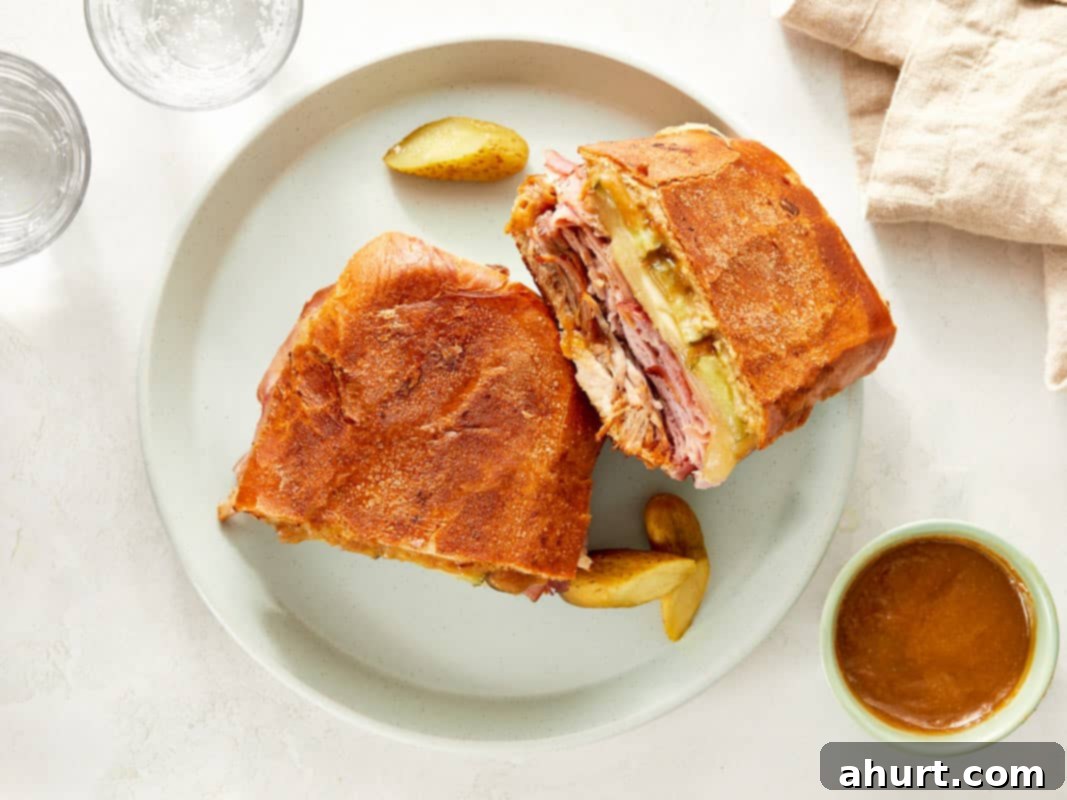 The Iconic Pressed Cuban 9 A perfectly pressed and halved Cuban sandwich served on a plate with additional pickles, showcasing its delicious layers and crispy exterior.