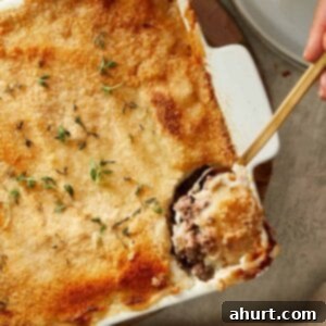 Spoonful of Shepherd’s Pie being served from a white baking dish, showing beef filling and mashed potato topping, garnished with fresh herbs.
