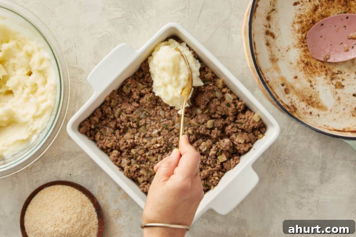 Creamy mashed potatoes being carefully and smoothly spread over the savory meat filling in a baking dish, creating the iconic top layer of the pie.