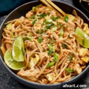 A vibrant bowl of homemade Pad Thai, featuring perfectly stir-fried rice noodles, succulent chicken pieces, golden tofu cubes, crunchy bean sprouts, and scrambled egg, all generously topped with crushed peanuts, fresh lime wedges, and bright green onions. Chopsticks rest alongside, inviting you to dig in.