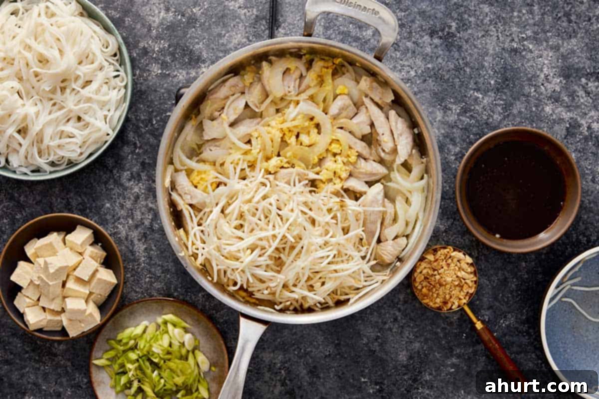Scrambled eggs cooking on one side of a wok, with cooked chicken and onions pushed to the other side, preparing to be combined for Pad Thai.