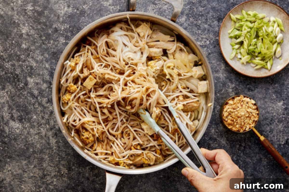 Cooked rice noodles being tossed with chicken, tofu, scrambled eggs, and bean sprouts in a wok using tongs, ensuring the sauce coats all ingredients evenly.