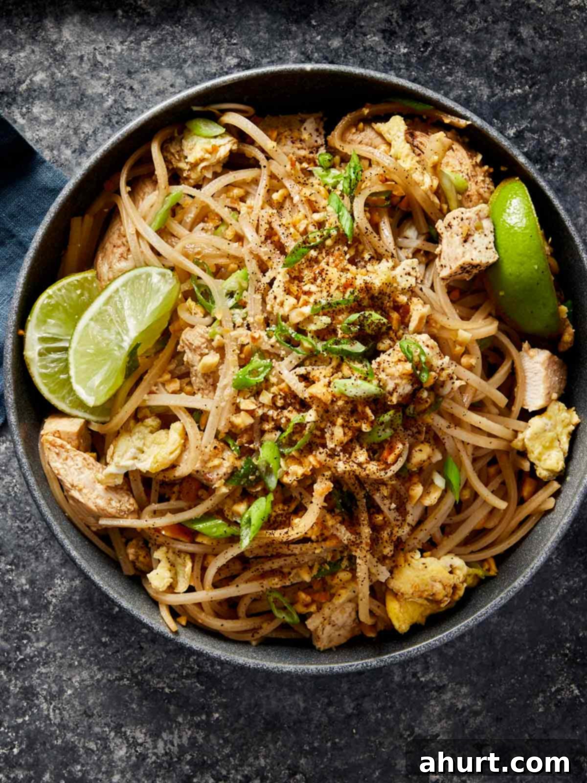 A top-down view of a generously portioned bowl of homemade chicken Pad Thai, elegantly arranged with scrambled egg, crisp bean sprouts, a sprinkle of crushed peanuts, fresh green onions, and bright lime wedges.
