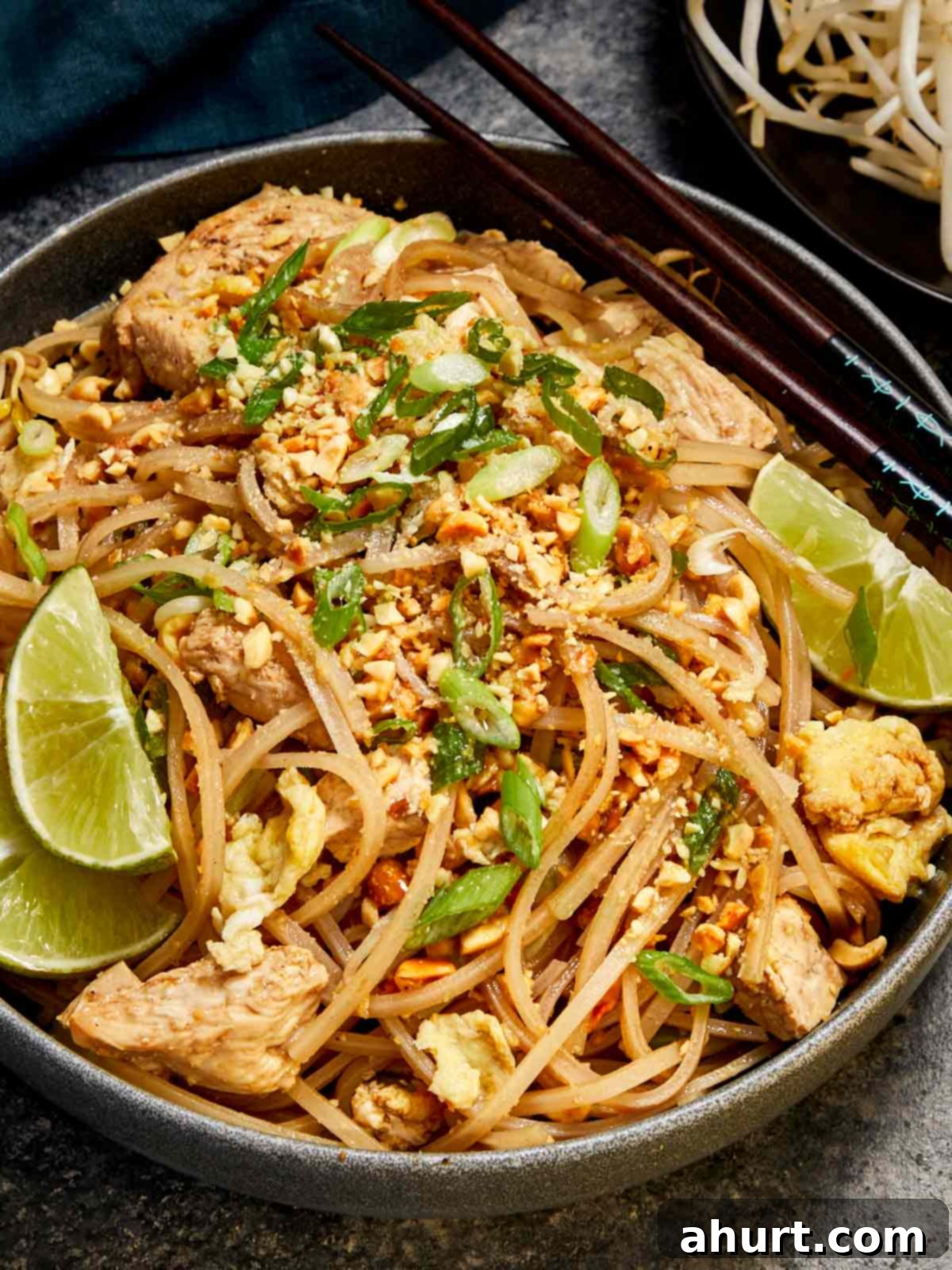 A beautifully presented bowl of chicken Pad Thai, showcasing stir-fried noodles, scrambled egg, bean sprouts, and crushed peanuts, finished with fresh lime wedges and green onions. The dish is accompanied by a pair of chopsticks, ready to be enjoyed.
