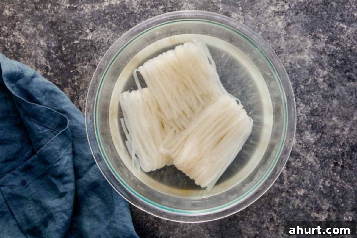 Uncooked flat rice noodles soaking in hot water inside a clear glass bowl, softening for Pad Thai preparation.