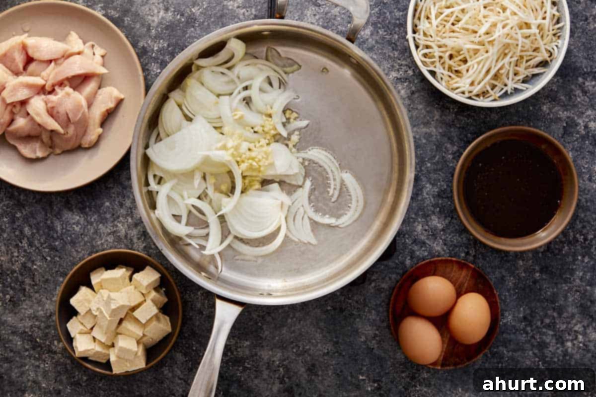 A wok containing sizzling vegetable oil, minced garlic, and thinly sliced onion, ready for stir-frying as the first step in making Pad Thai. Other ingredients like chicken, eggs, tofu, bean sprouts, and sauce are visible nearby.
