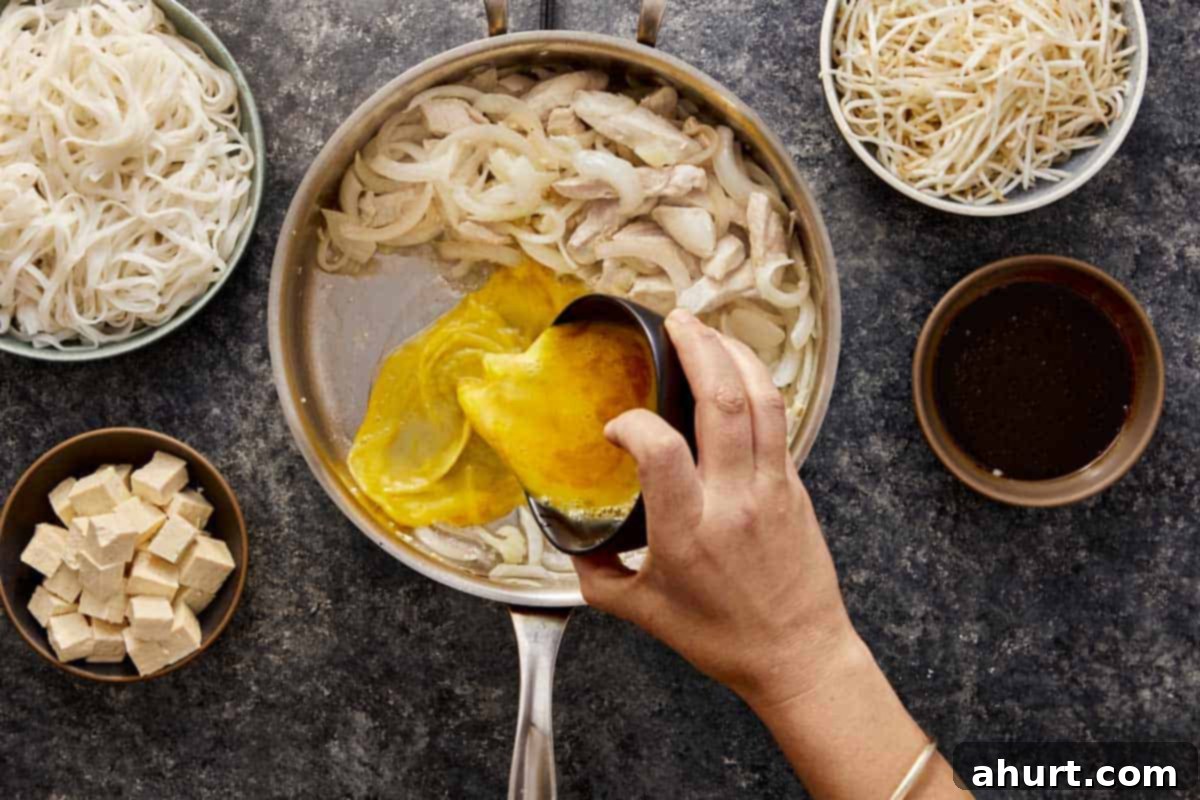 A hand pouring lightly whisked eggs into an empty section of a hot wok, next to already cooked chicken and onions, to create scrambled egg for Pad Thai.