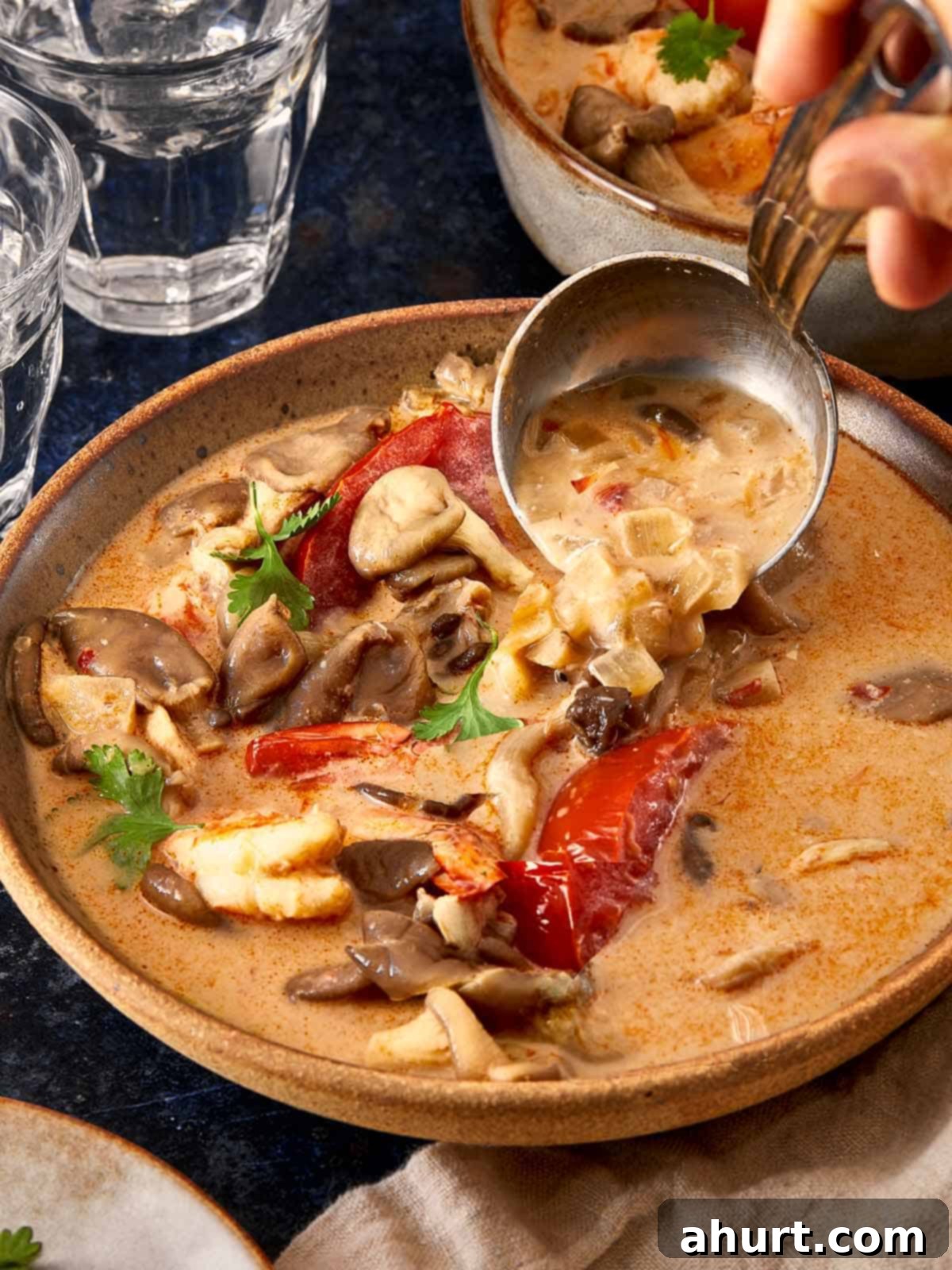 A warm bowl of Tom Yum Kha soup filled with shrimp, oyster mushrooms, tomatoes, and a creamy coconut broth being ladled into a bowl.