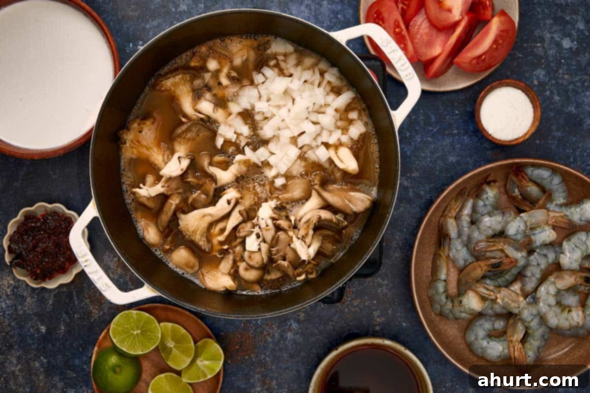 Pot with onions and oyster mushrooms simmering in broth, surrounded by shrimp, tomatoes, limes, and coconut milk.