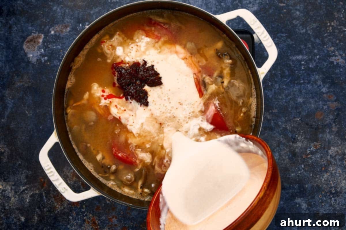 Pouring coconut milk and Thai chili paste into a pot of simmering soup.
