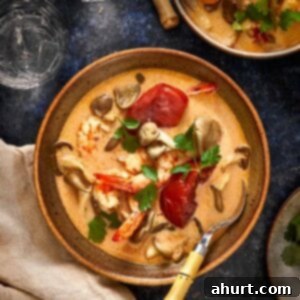 Overhead shot of creamy Thai Tom Yum Kha soup with shrimp, mushrooms, and tomatoes served in a rustic bowl with a bamboo-handled spoon and fresh cilantro on top.