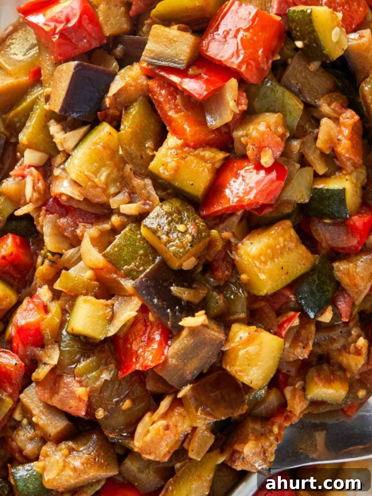A vibrant bowl of ratatouille, filled with assorted cooked vegetables, accompanied by a spoon for serving.