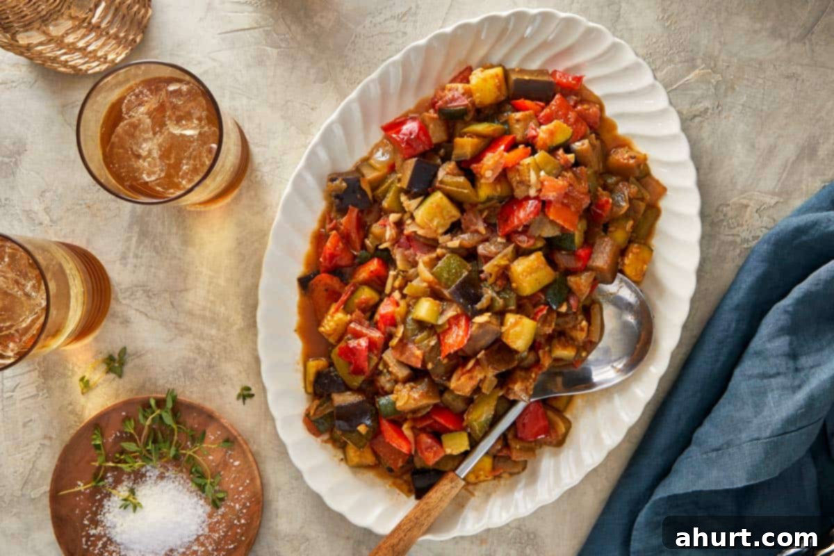 Ratatouille served on an oval platter with a spoon, surrounded by drinks and herbs.