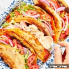 Close-up of viral smash burger tacos in a blue speckled tray, filled with melted cheese, crisp lettuce, fresh tomato, red onion, and topped with creamy sauce.