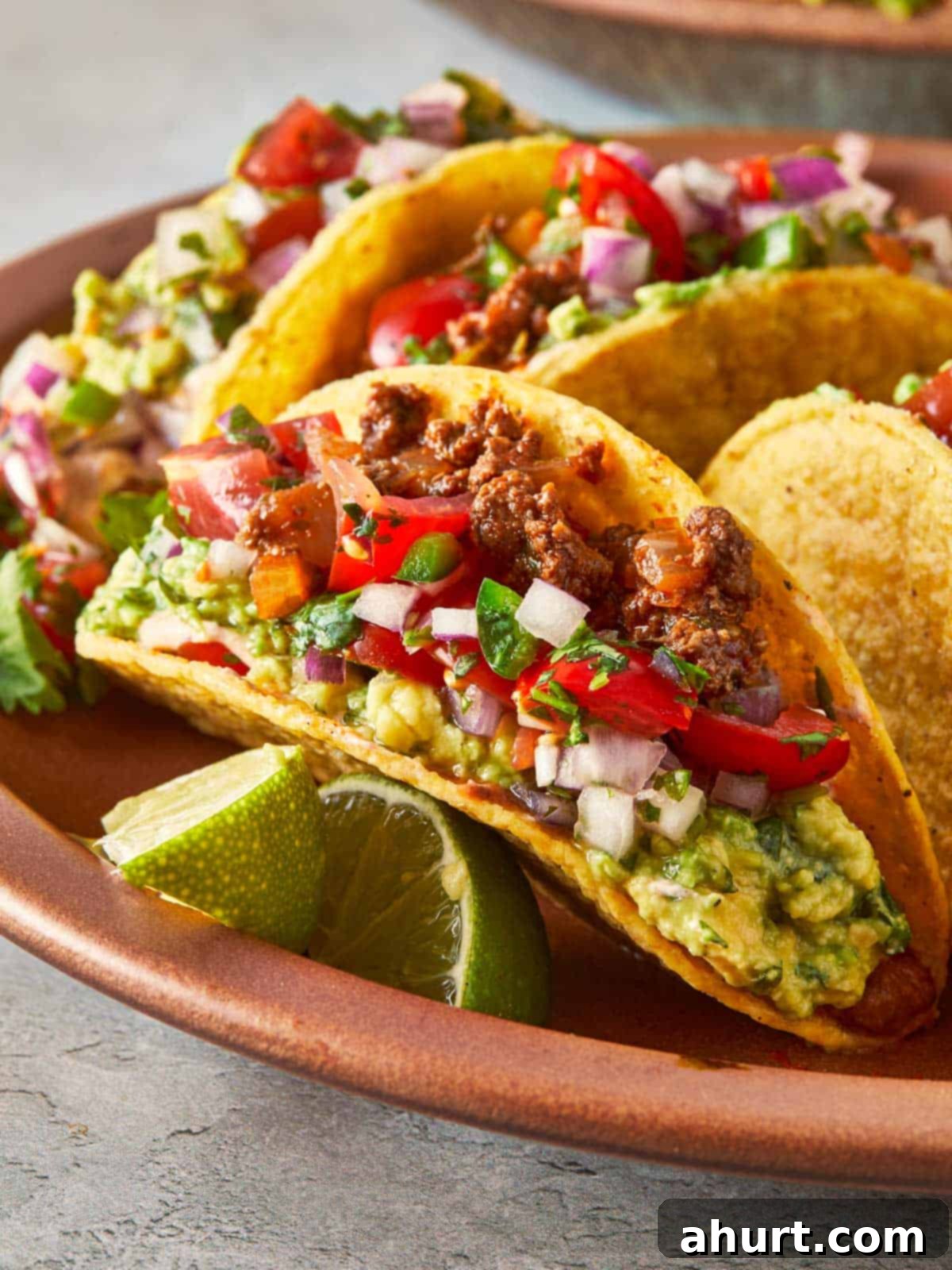Close-up of a beef taco with guacamole, salsa, and lime wedges on a plate, showcasing its delicious layers and vibrant toppings.
