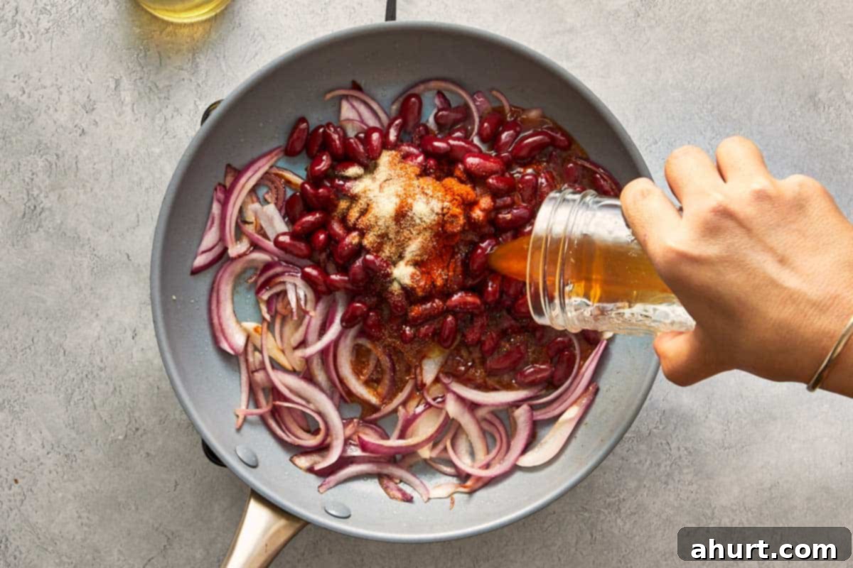 Red beans and thinly sliced red onions cooking together in a pan with aromatic spices, forming a flavorful base.