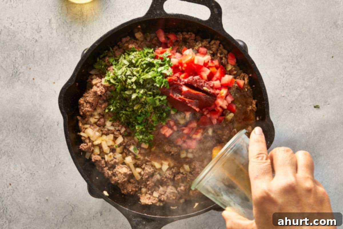 Cooked ground beef mixture simmering in a skillet with diced tomatoes, tomato paste, and fresh cilantro, creating a rich, saucy texture.