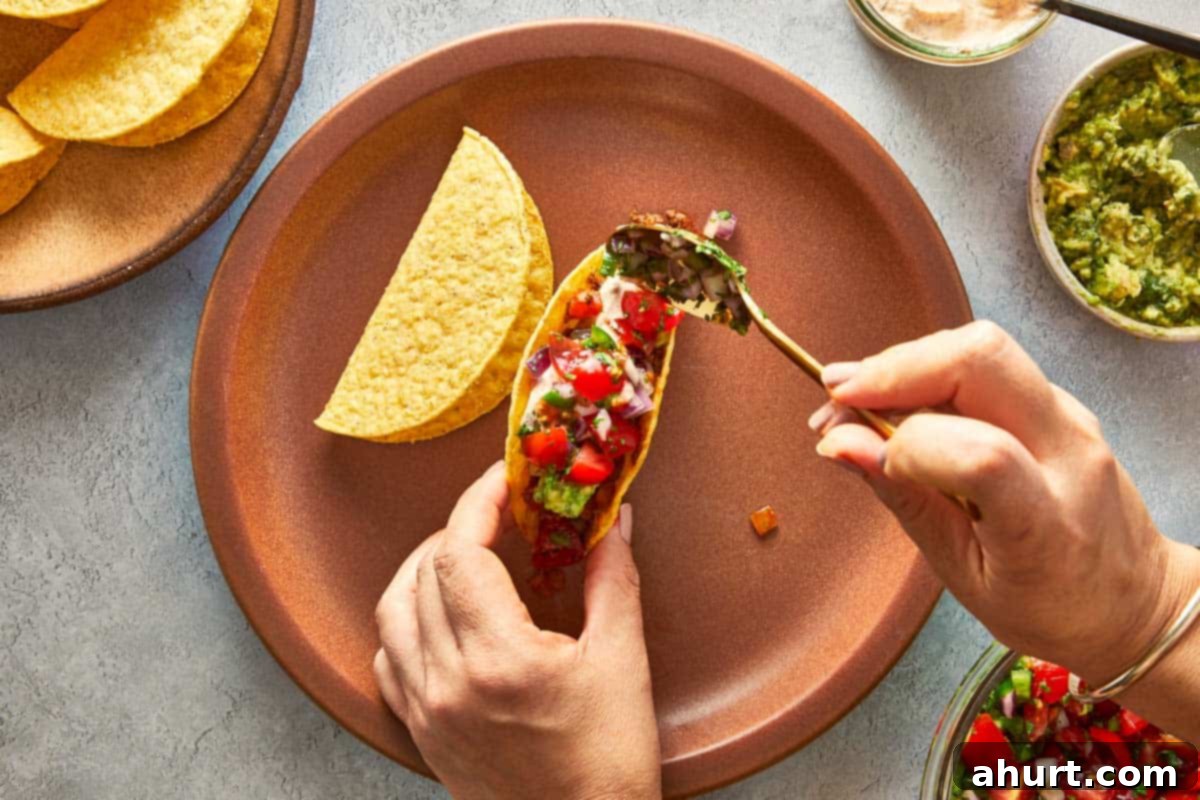 Hands meticulously assembling a taco, filling a shell with seasoned beef, creamy guacamole, and fresh pico de gallo.