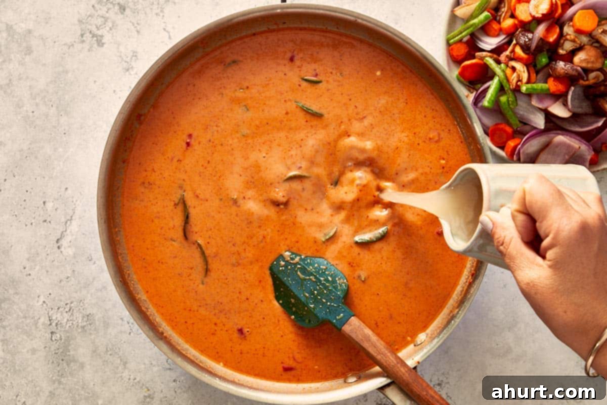 Coconut milk and chicken stock being poured into the curry base, creating a creamy orange sauce with floating lime leaves.