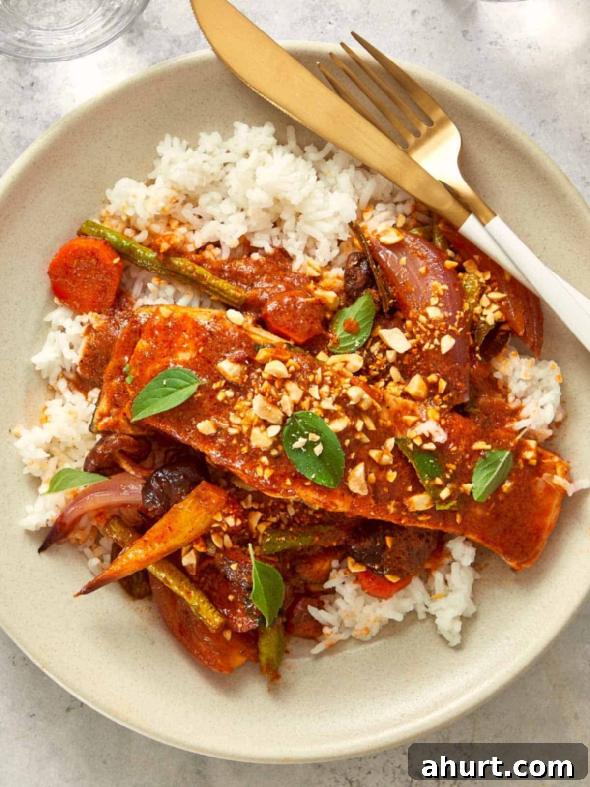 Overhead shot of Salmon Panang Curry served over jasmine rice, garnished with fresh Thai basil and chopped roasted peanuts. Tender salmon fillets are coated in a rich red curry sauce, surrounded by colorful vegetables, creating a vibrant and appealing dish.