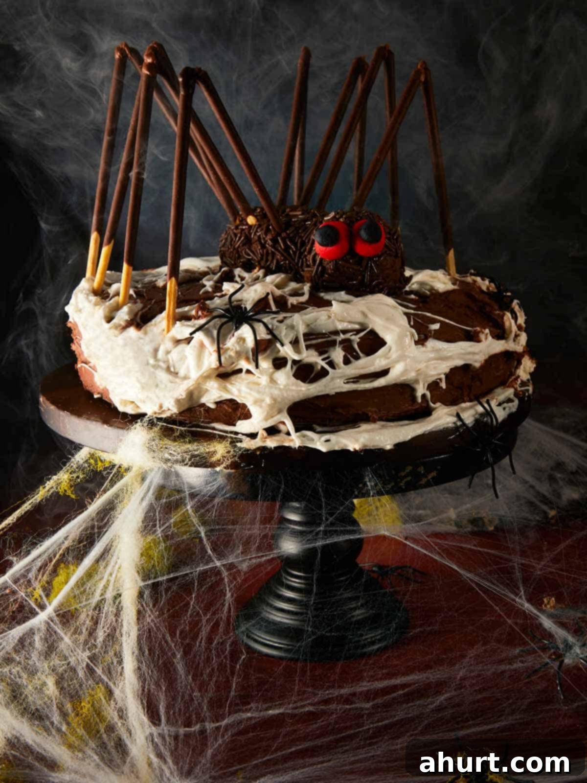 Halloween chocolate layer cake styled as a giant spider, surrounded by fake cobwebs and plastic spiders on a dark red table.