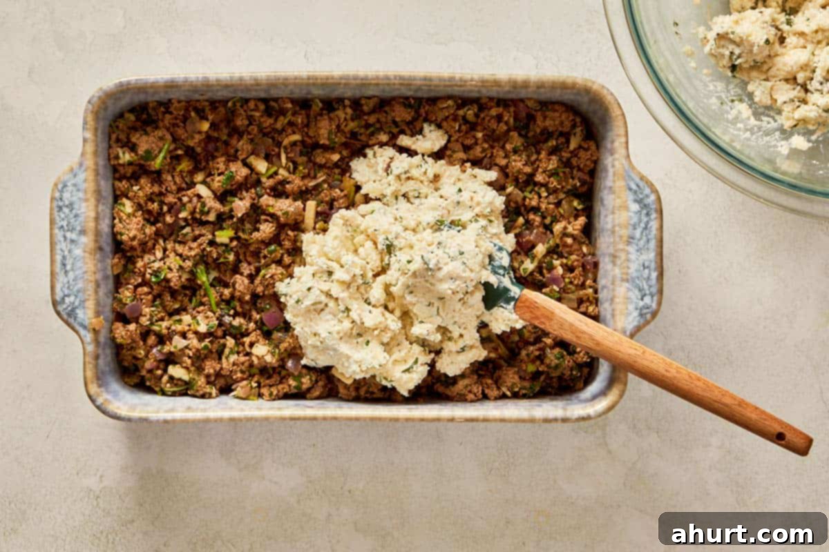 Spreading cheese mixture over cooked beef in a baking dish.