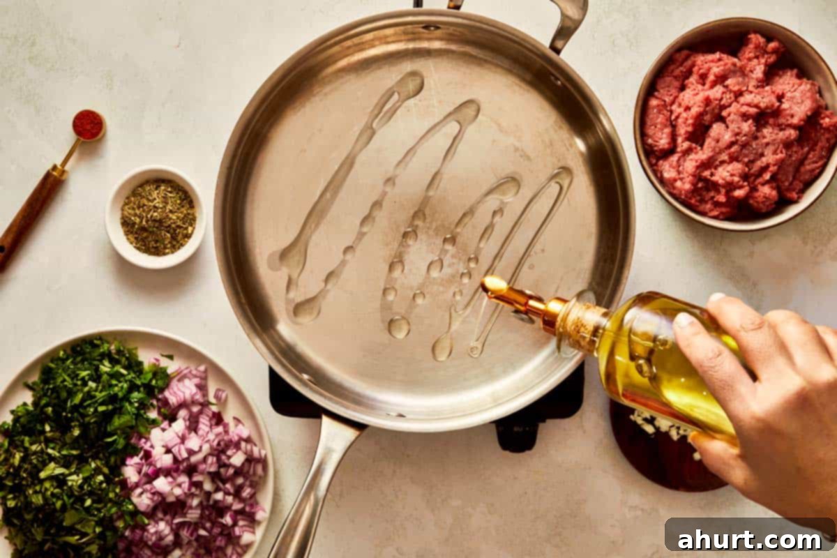 Drizzling olive oil into a hot pan with beef and herbs nearby.