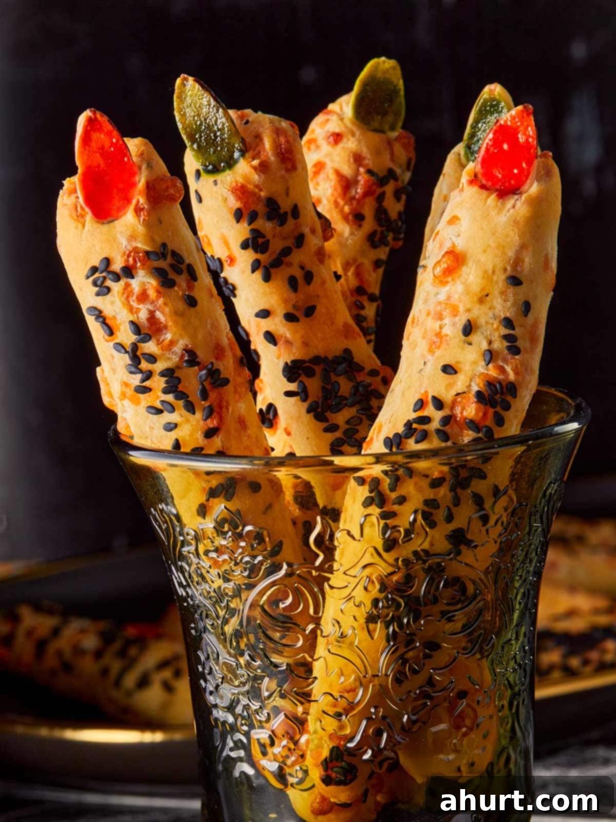Close-up of Witch Finger Grissini breadsticks standing in a decorative glass, topped with red and green almond “nails” and coated with black sesame seeds for a spooky Halloween look.