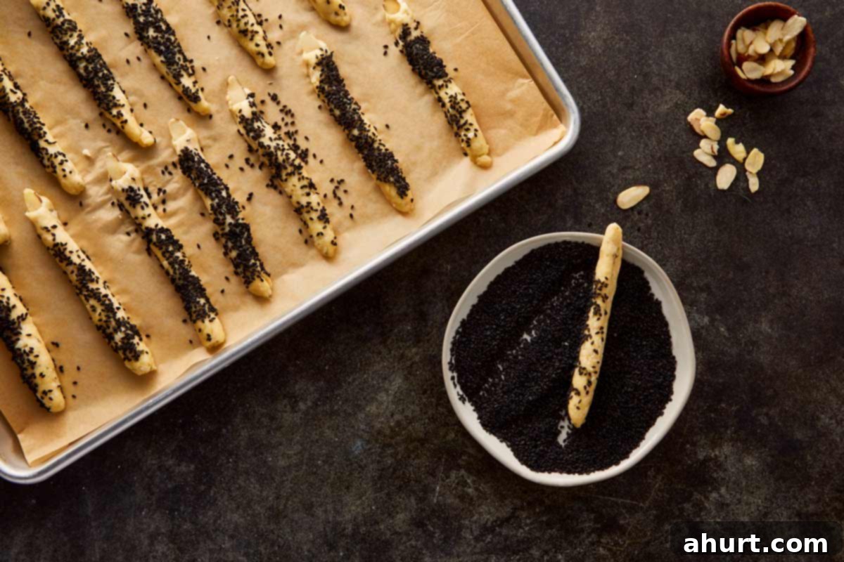 Fingers of dough being coated with black sesame seeds on a parchment-lined tray for texture and color.