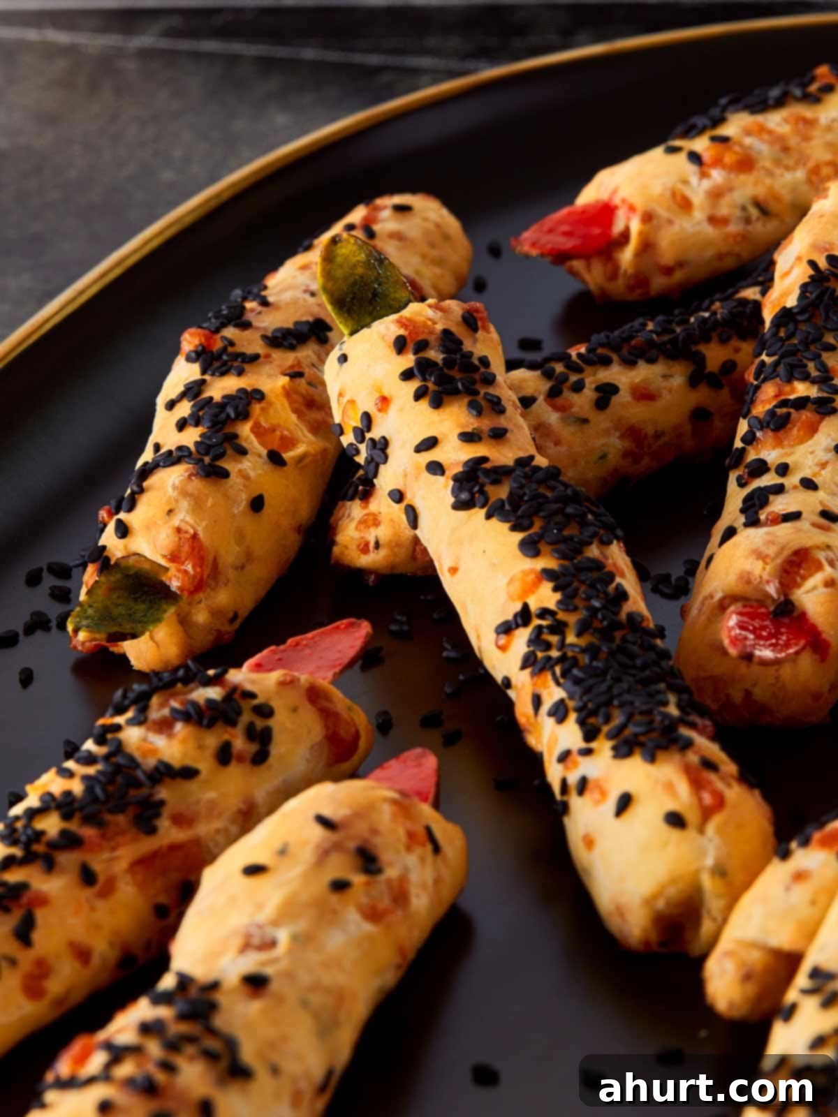 Close-up of freshly baked Witch Finger Grissini on a dark serving plate, coated with black sesame seeds and topped with red and green almond “nails” for a spooky Halloween look.