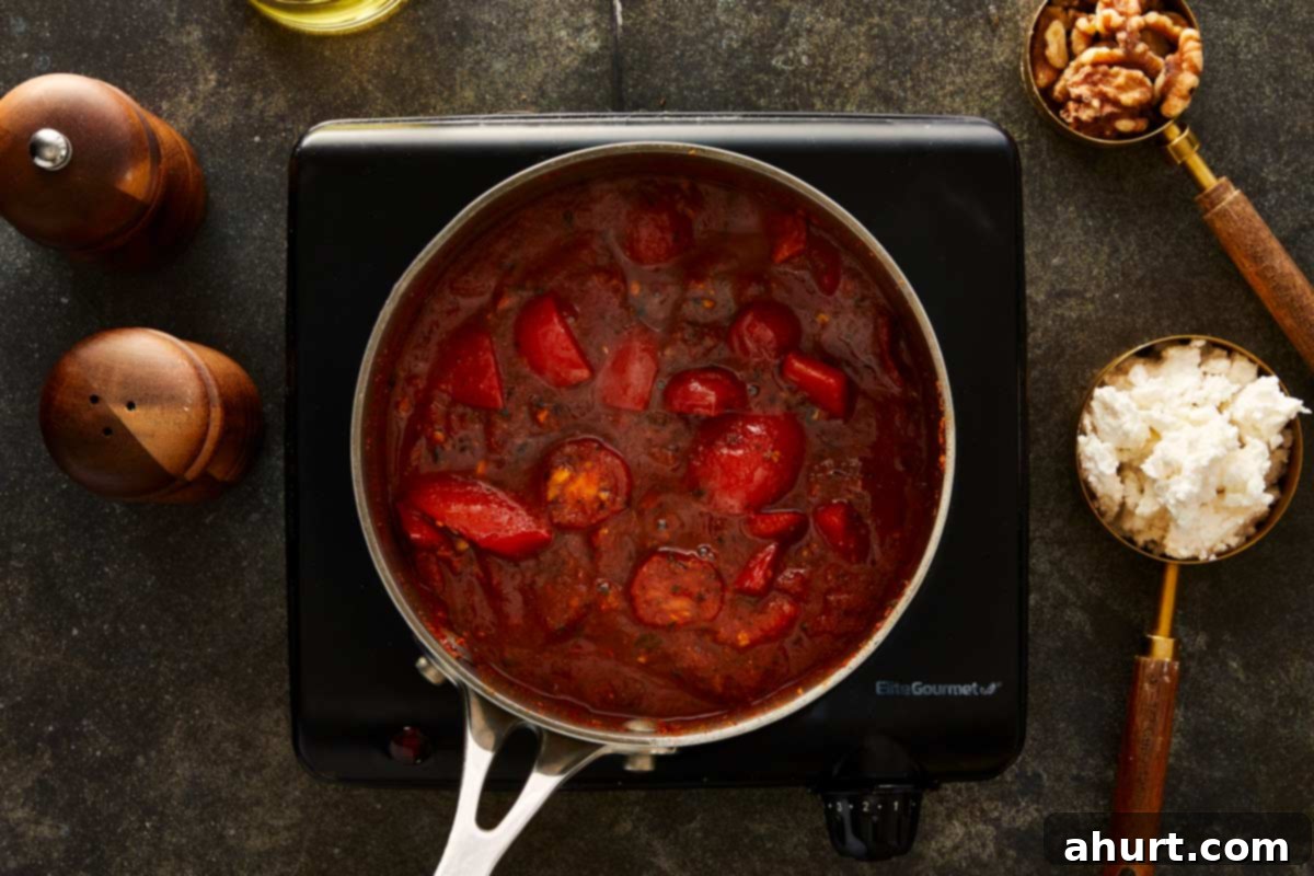 Simmering mixture of tomatoes and red peppers in a saucepan, thickened into a rich red sauce, with feta and walnuts on the side.
