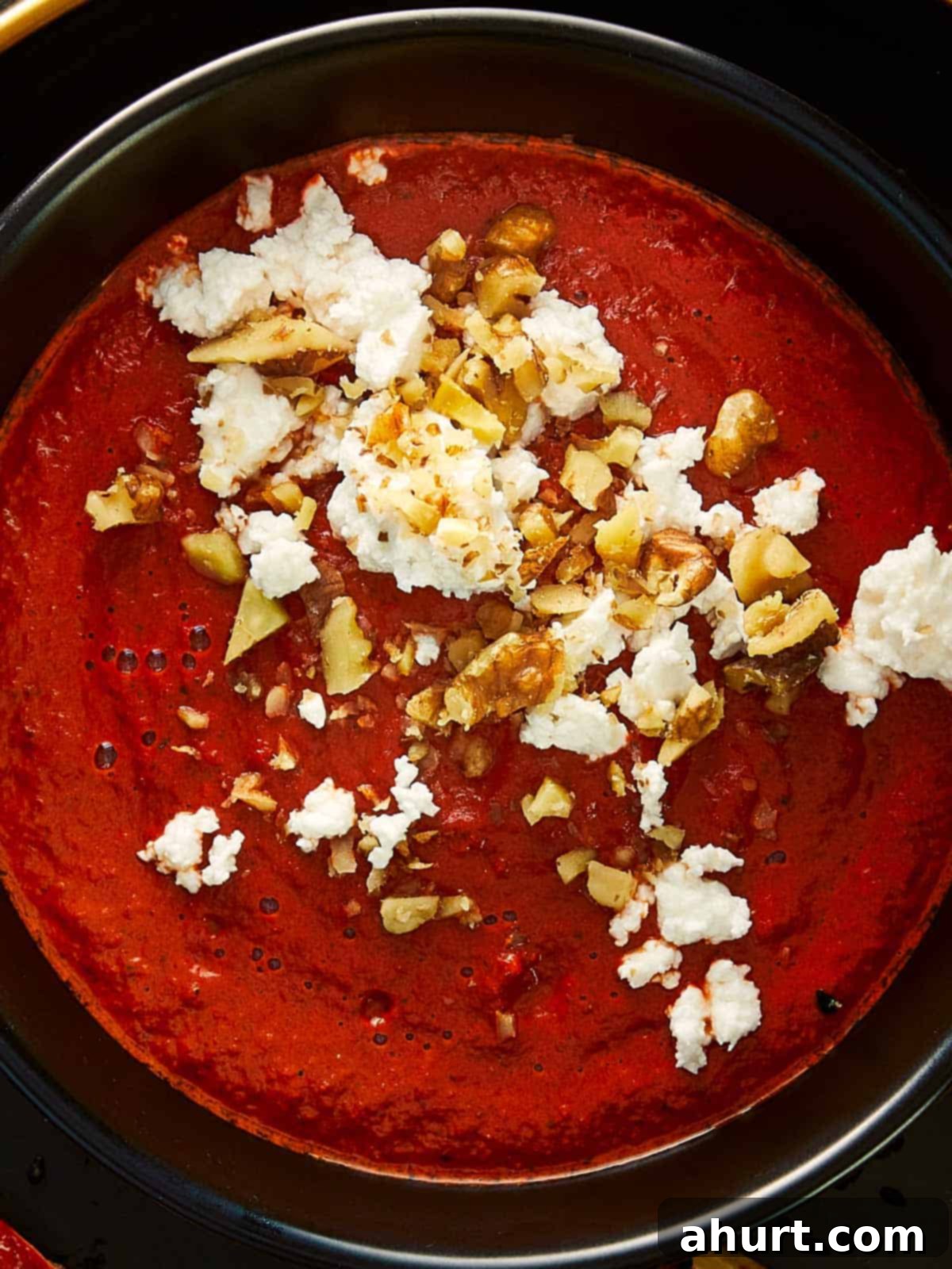 Close-up of a rich, vibrant red dip topped with crumbled feta cheese and chopped walnuts, served in a black bowl. The creamy, smoky texture and contrast of colors give this Bloody Dip an appetizing Halloween vibe.