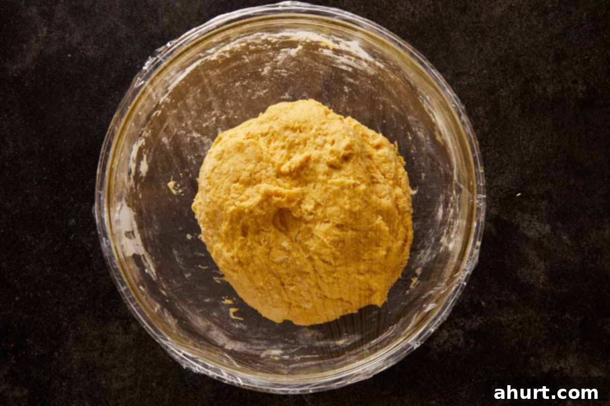 Smooth pumpkin bread dough resting in a glass bowl, covered with plastic wrap and ready to rise.