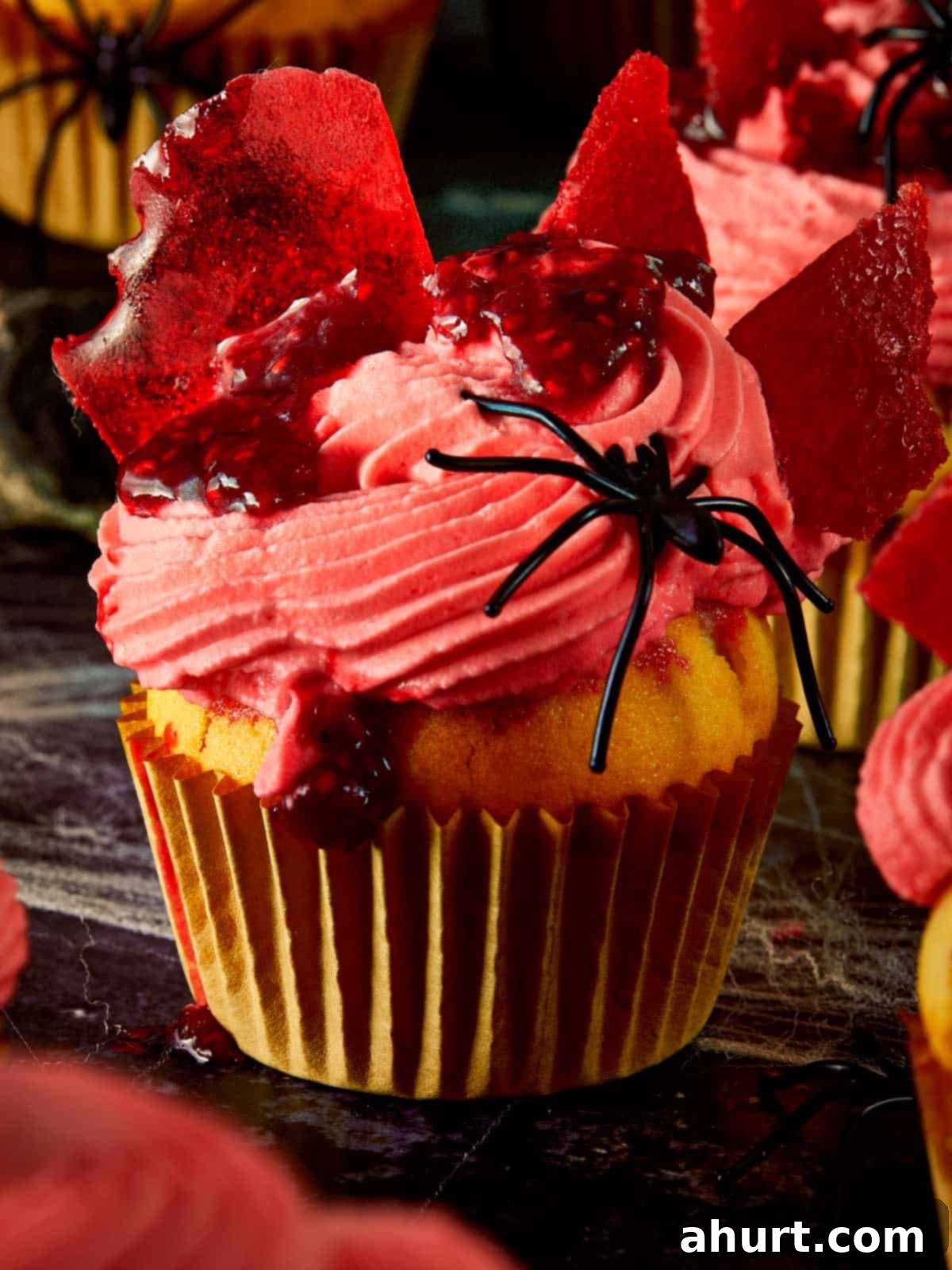 Close-up of a Halloween raspberry muffin topped with pink whipped cream, raspberry coulis, and broken glass candy shards, decorated with fake black spiders. Perfect for spooky dessert ideas.