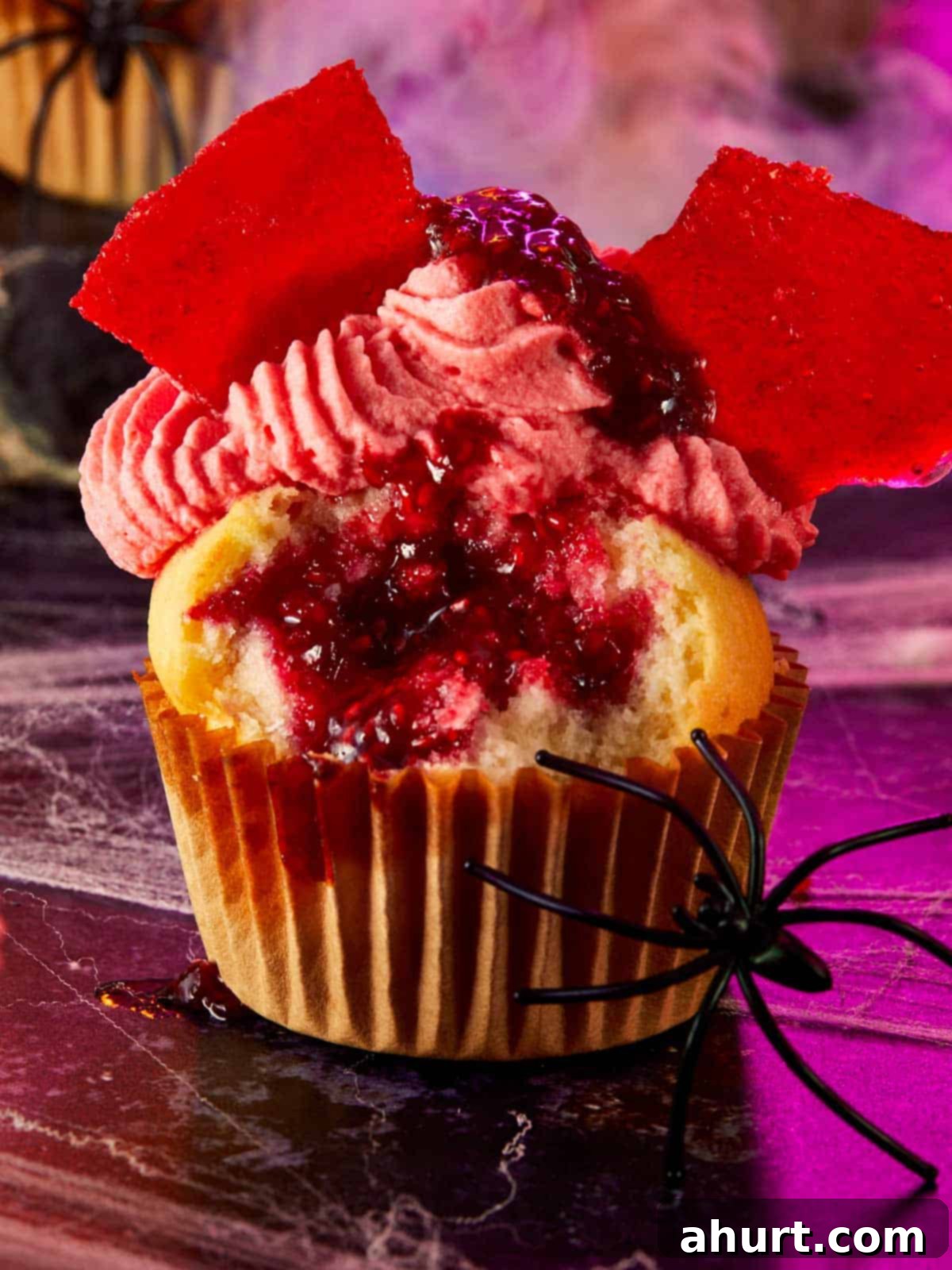 Raspberry muffin cut open to reveal a red raspberry filling, topped with whipped cream, candy glass shards, and a fake black spider on a spooky purple surface. A delicious Halloween treat.