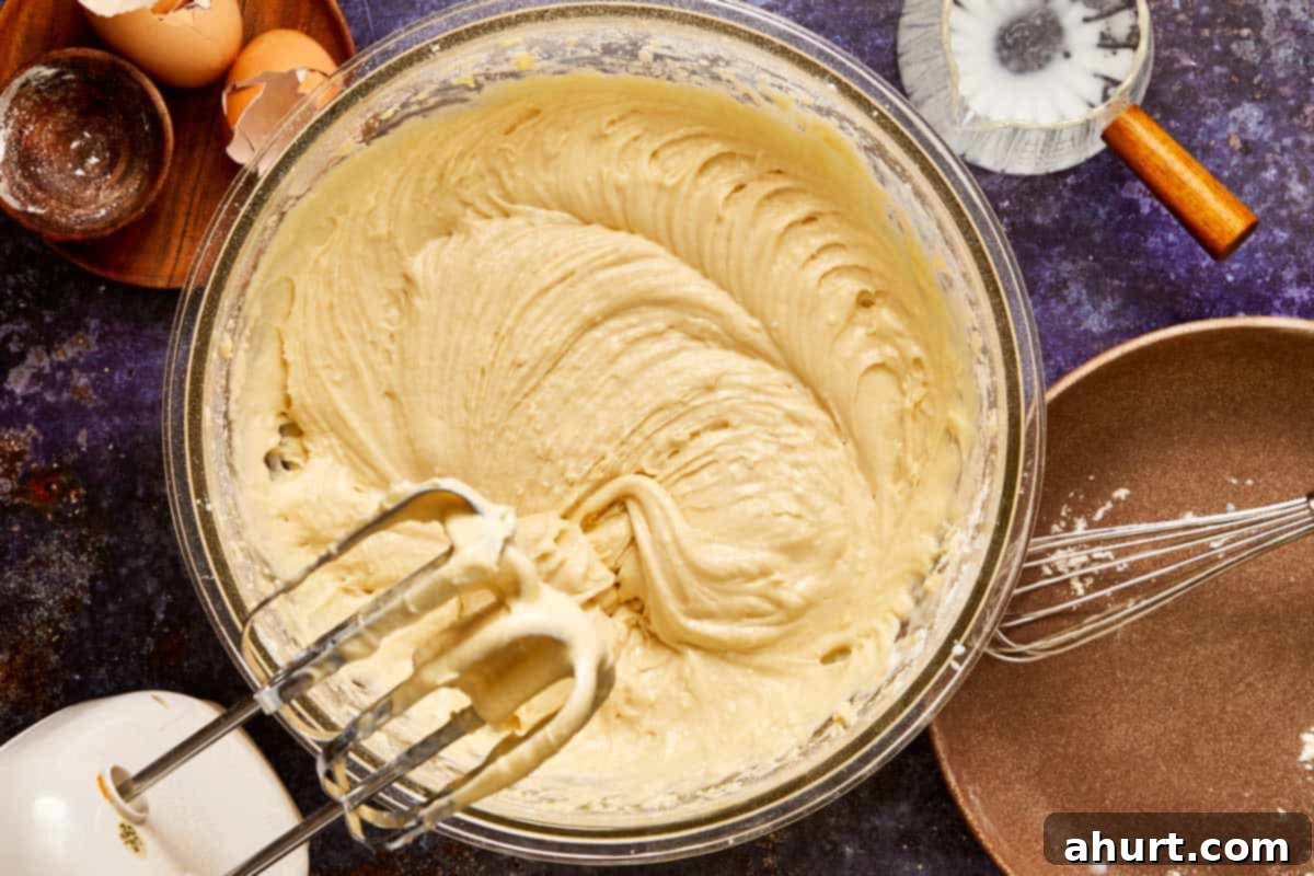 Mixing bowl filled with smooth muffin batter being whipped with an electric mixer, surrounded by eggs and baking tools. Achieving the perfect vanilla muffin texture.