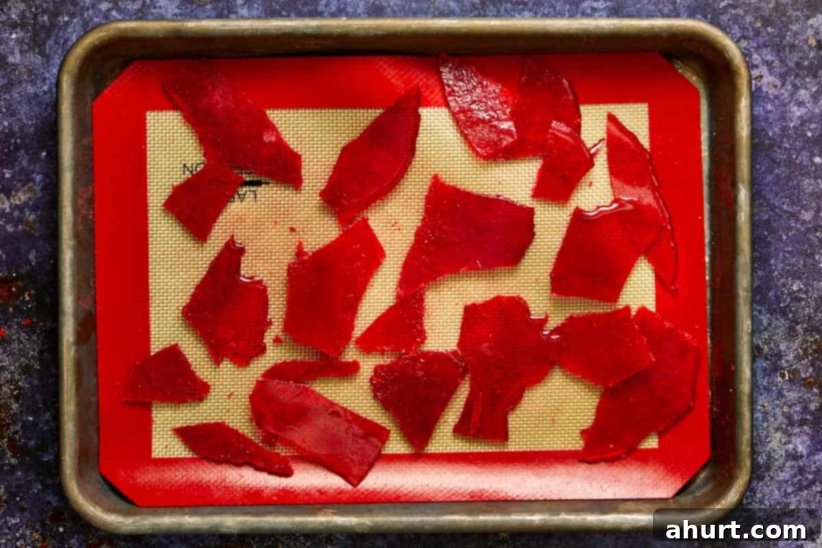 Tray of bright red caramel shards resembling broken glass cooling on a silicone mat, ready for decoration. Adds a spooky crunch to Bloody Raspberry Muffins.