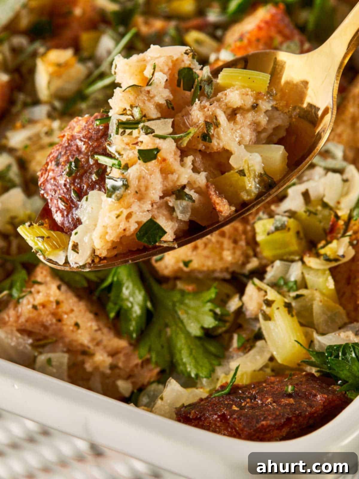 Classic Buttery Herb Stuffing 2 Golden cubes of buttery herb stuffing in a white baking dish, topped with fresh parsley, sage, and rosemary. A gold spoon lifts a perfect bite showing the soft center and crisp edges.