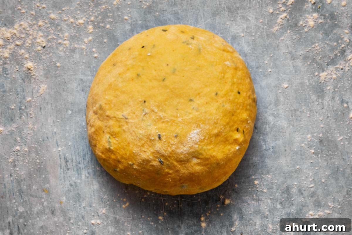 A perfectly shaped ball of sweet potato bread dough resting in a lightly oiled glass bowl, covered with plastic wrap, indicating the first rise
