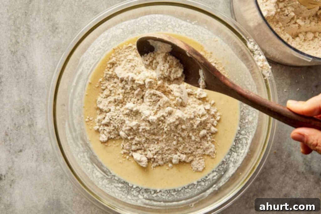 a top down image of a glass mixing bowl with white batter in the bottom of the bowl and white powder on top getting ready to be mixed with a wooden mixing spoon