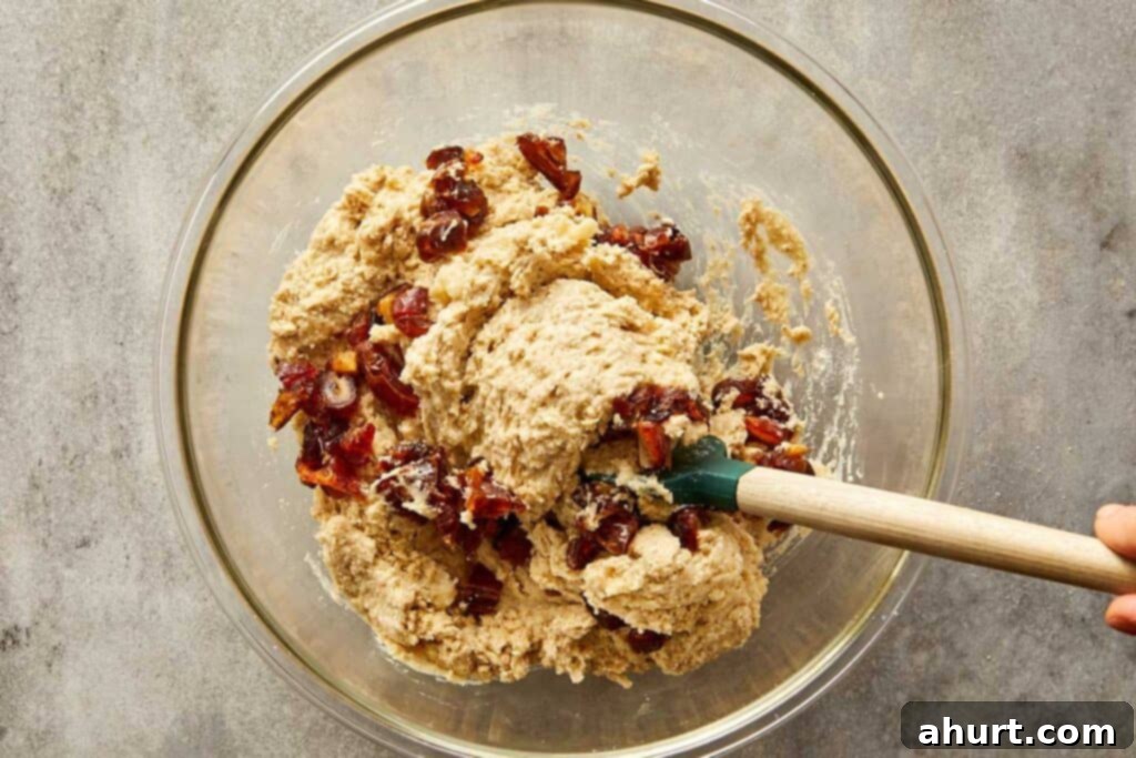 a top down image of a glass mixing bowl with white batter in the bottom of the bowl and white powder on top getting ready to be mixed with a wooden mixing spoon with fruit inside