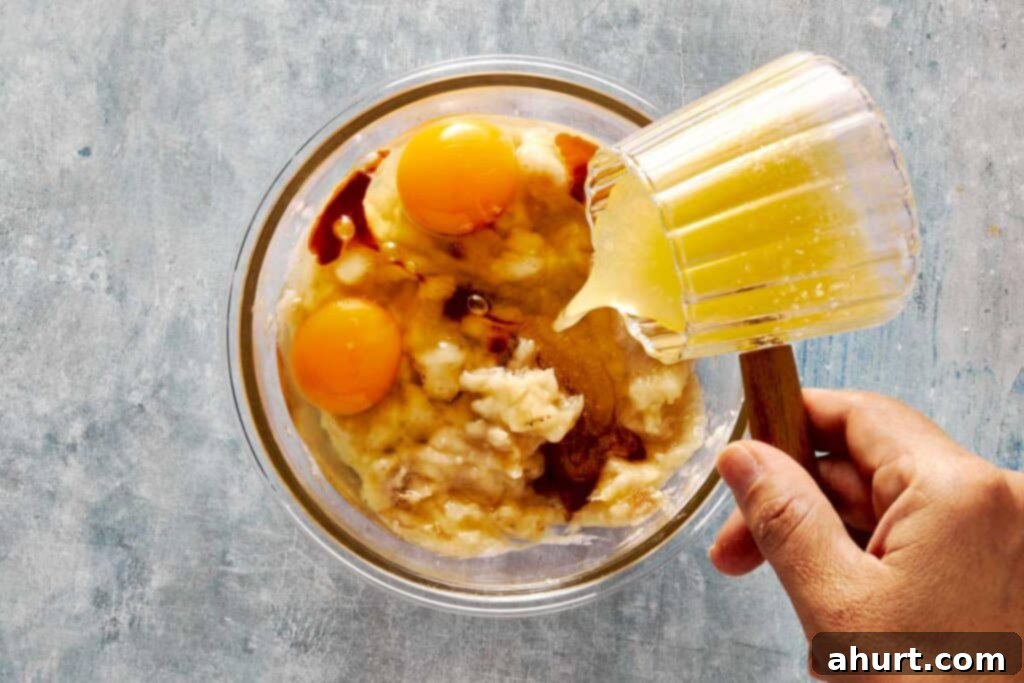 A top-down view of wet ingredients being gently mixed into the dry ingredients in a glass bowl, with a glass jug pouring more liquid into the mixture.