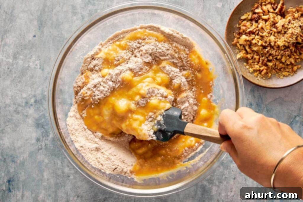 A top-down view of a green spatula gently mixing the wet and dry ingredients together in a large glass bowl sitting on a countertop.